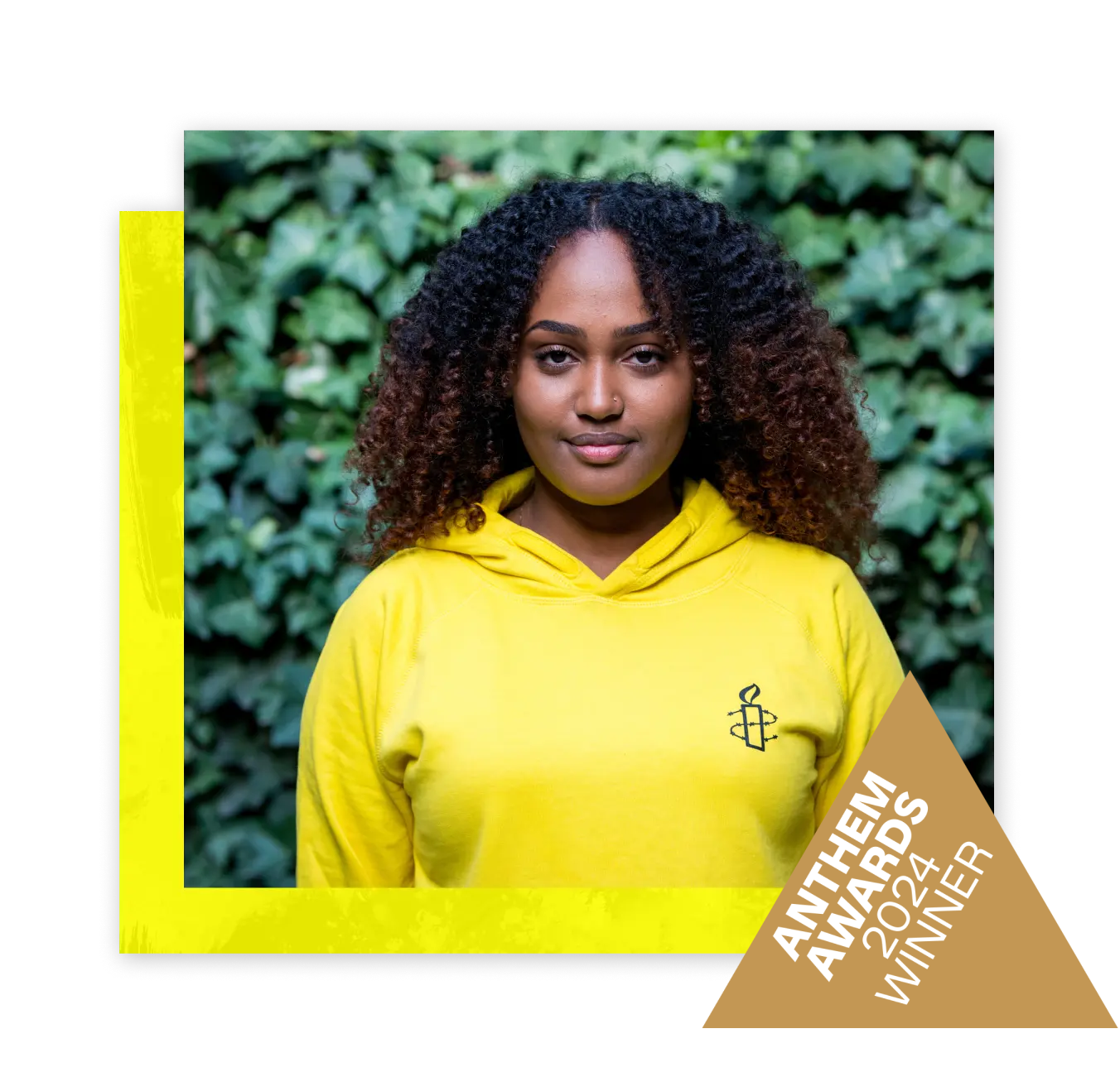 A woman in a bright yellow t-shirt with the Amnesty International logo on it standing in front of a lush green, leafy background. The photo is overlapping a bright yellow square and there is a small badge in the bottom corner that reads 